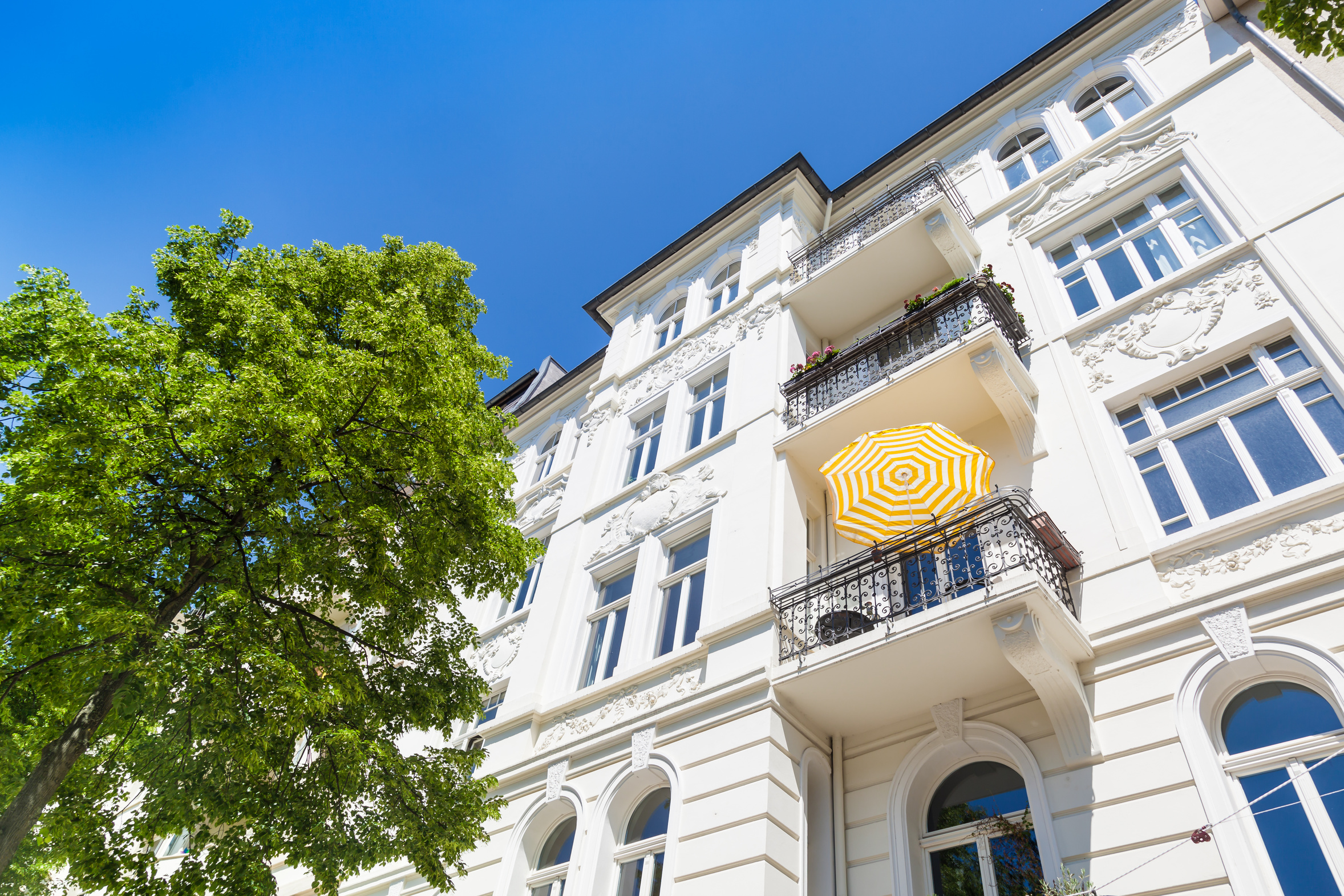 Helle Altbaufassade mit Balkonen und grünem Baum